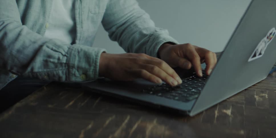 A man smiles as he types on a laptop sitting at a wooden table near a window