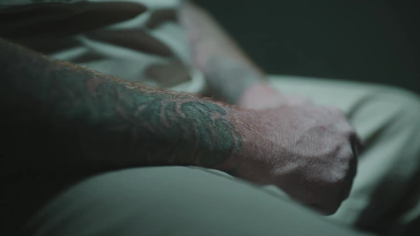 A man wearing a prison uniform sitting clenching his hands with tattoos on both arms