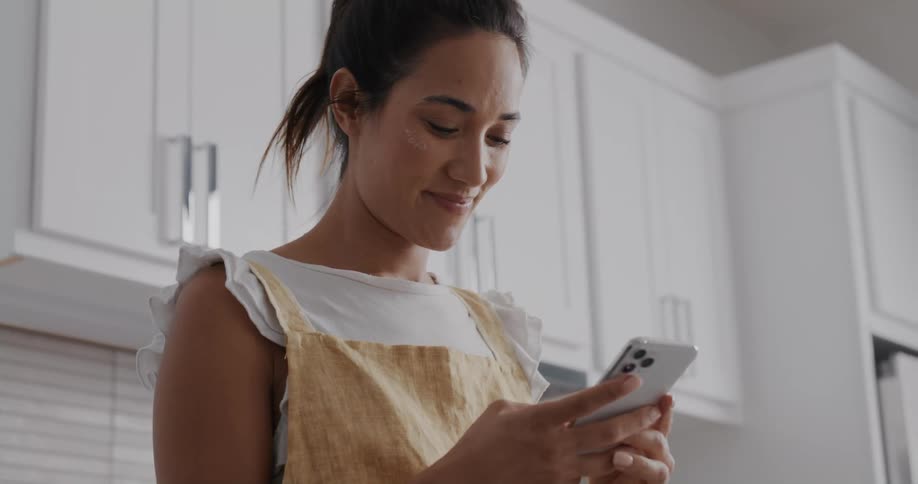 A woman is texting on her cellphone and standing in the kitchen