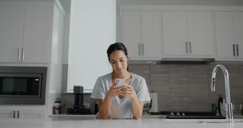 A woman is standing in her kitchen and texting on her cell phone