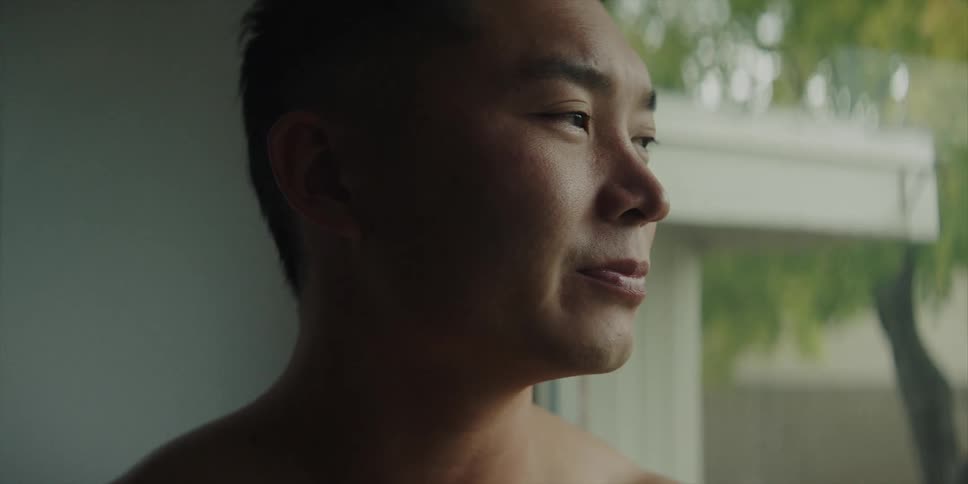 Topless muscular man drinks from gray cup smiling looking out the window