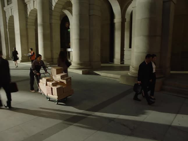Pedestrians walk down a city sidewalk while a woman pushing a cart with packages looks at her watch and walks quickly past them
