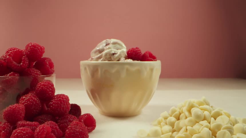 A bowl of ice cream with raspberries next to fresh raspberries and a pile of white chocolate chips