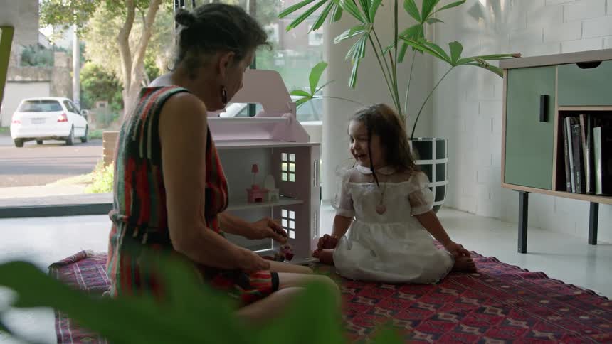 A little girl sits on the floor in a home and plays with her dollhouse with her grandmother