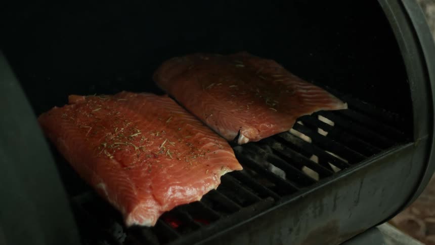 A hand lifts a charcoal grill lid displaying the two salmon fillets cooking