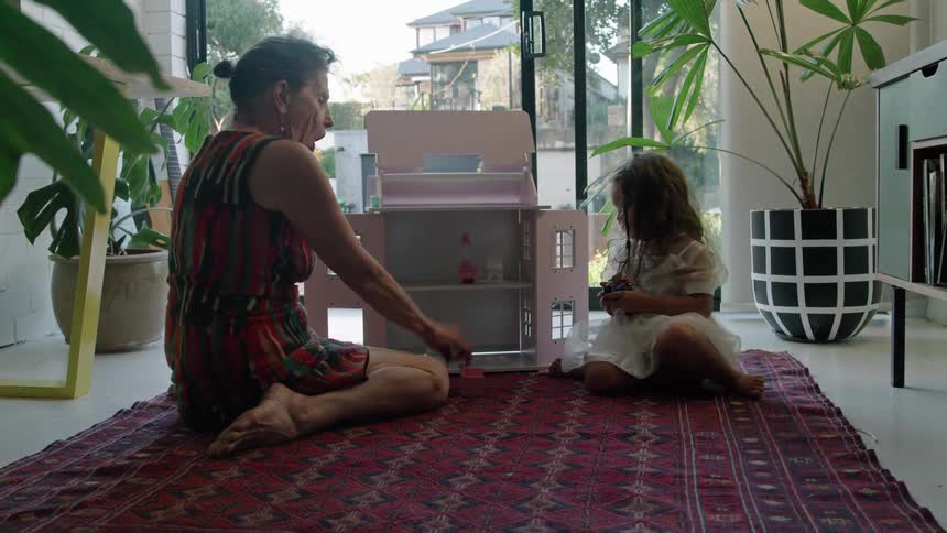 A woman is sitting on the floor playing with dolls and a doll house with her granddaughter