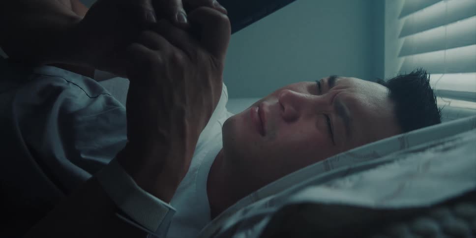 Sleepy man lying in bed uses smartphone next to window blinds in the morning