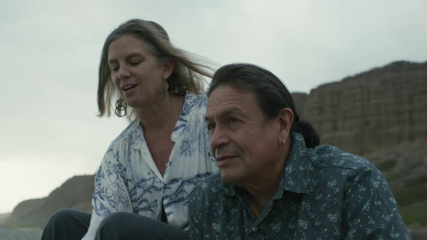 Man with long hair and blue shirt and blonde woman with white shirt sit on pebble beach on cloudy day