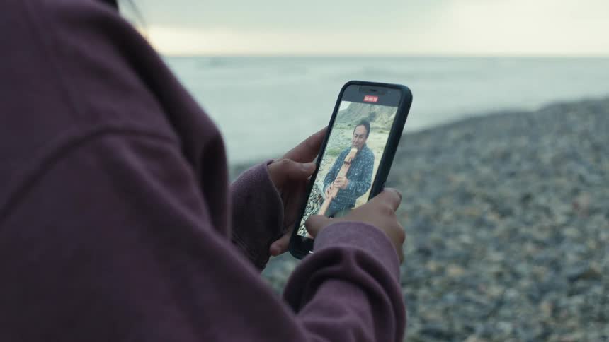 A woman holding a smart phone filming a man playing a native american drone flute on the beach