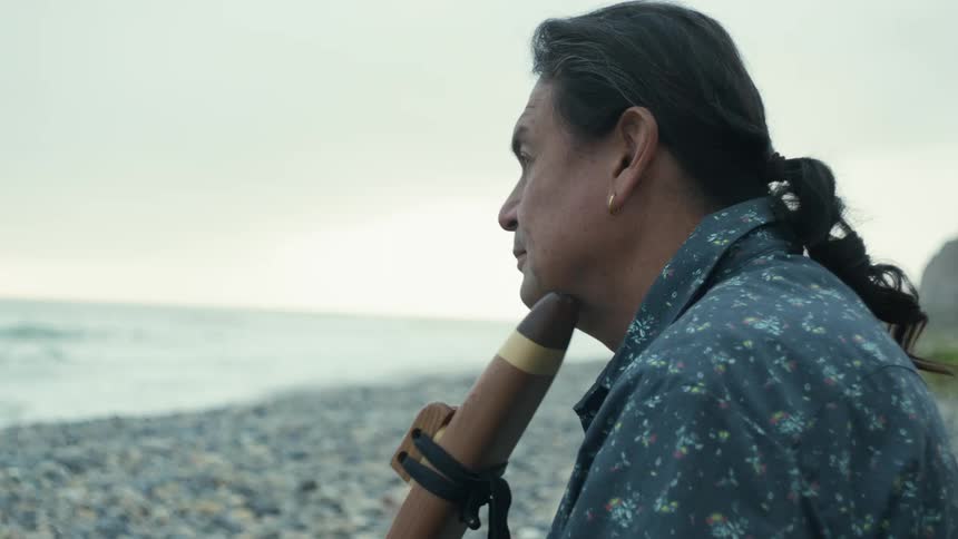 A native american man caresses a wooden drone flute then lifts it to play at the beach