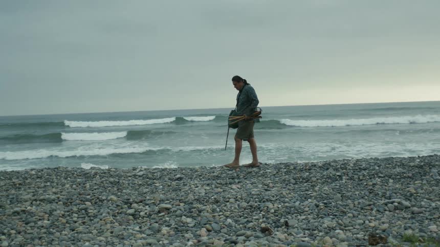 A man walking along a rocky beach stops to pick up something near the waves
