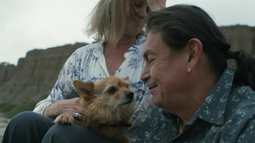 A couple is sitting on a rocky beach near cliffs with their small dog