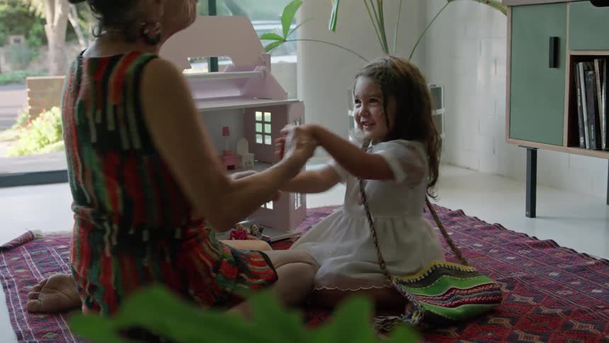 Old woman with striped dress plays with toddler girl with wavy hair and white dress next to pink model house sitting on red carpet in living room at home