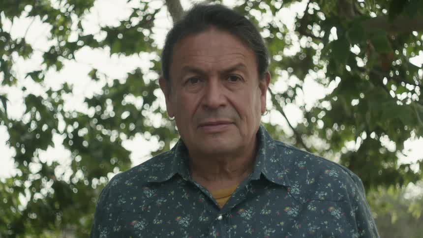Man with blue shirt and long hair looks at camera under tree