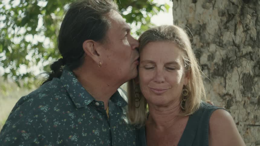 A man and woman stand under a tree and he rubs her back and kisses her on the forehead