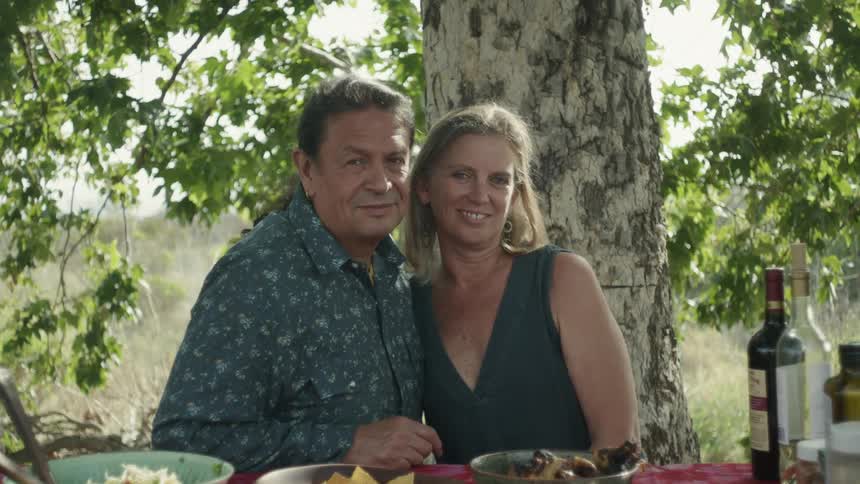 A man pulls a woman close to him as they sit at a table outside with food and wine