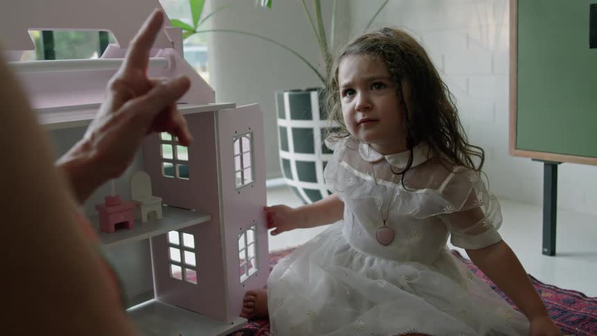 Old woman with striped dress plays with toddler girl with wavy hair and white dress next to pink model house sitting on red carpet in living room at home