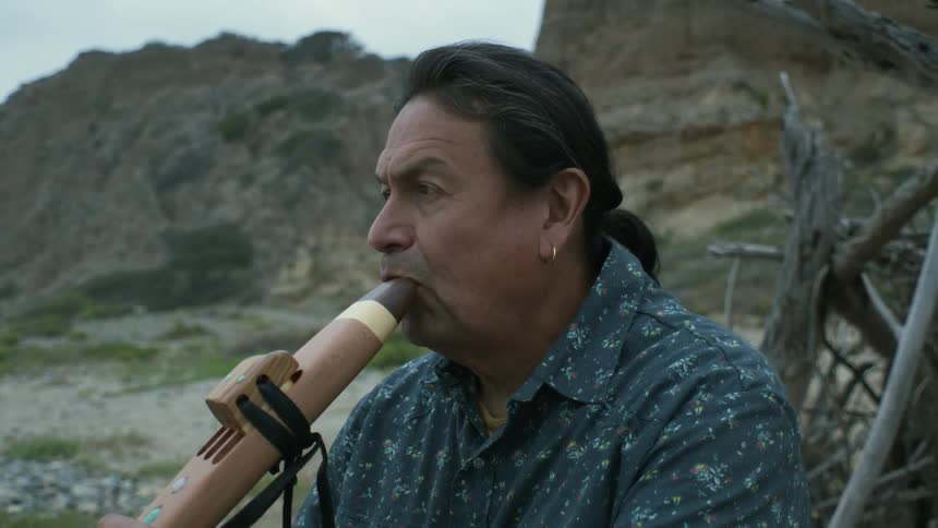A man sitting on driftwood at the beach begins to play a native american drone flute