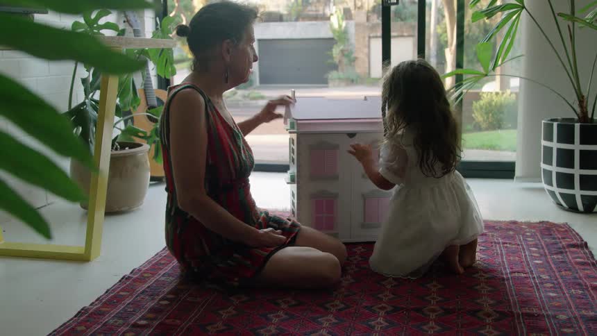 A woman is sitting on the floor playing with a doll house with her granddaughter and she gives her a big hug