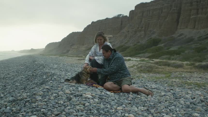 A couple is relaxing on a rocky beach with their dog