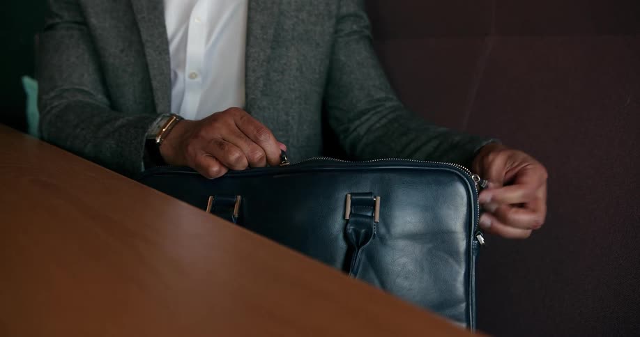 A man dressed in a suit and seated at a table puts a laptop into a briefcase and zips it