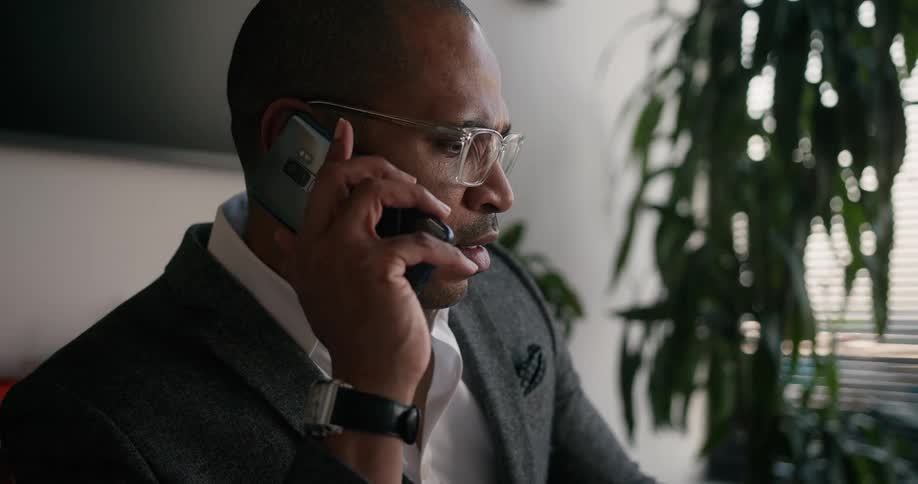 Black man sitting at the desk in his office talking on the phone while using his laptop