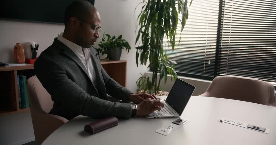 A man is sitting at a table in an office and he puts on his glasses and types on a laptop computer