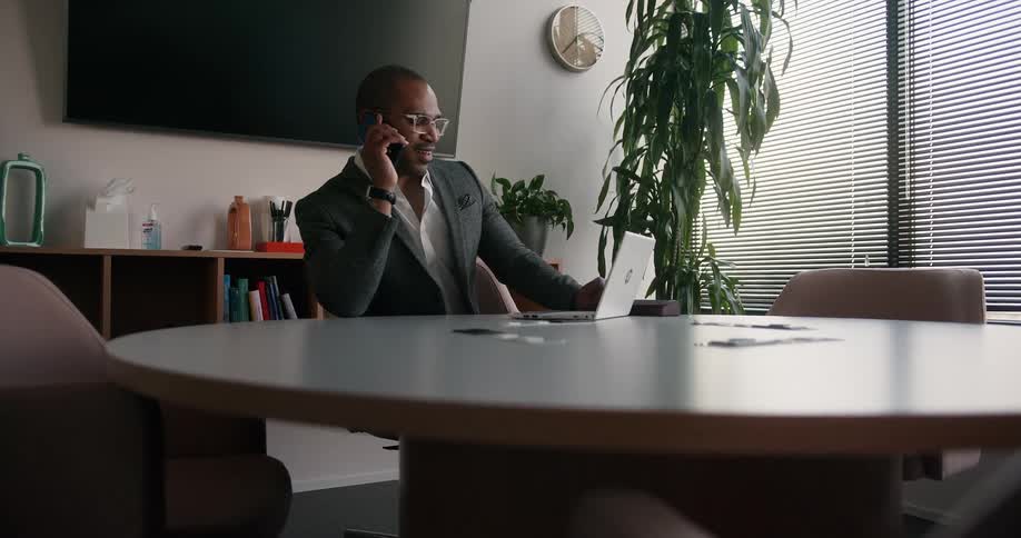 A man is sitting at a table with a laptop computer as he talks on his phone