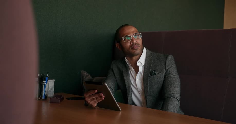 A man is sitting at a table holding a tablet computer and looking at it and then looking up