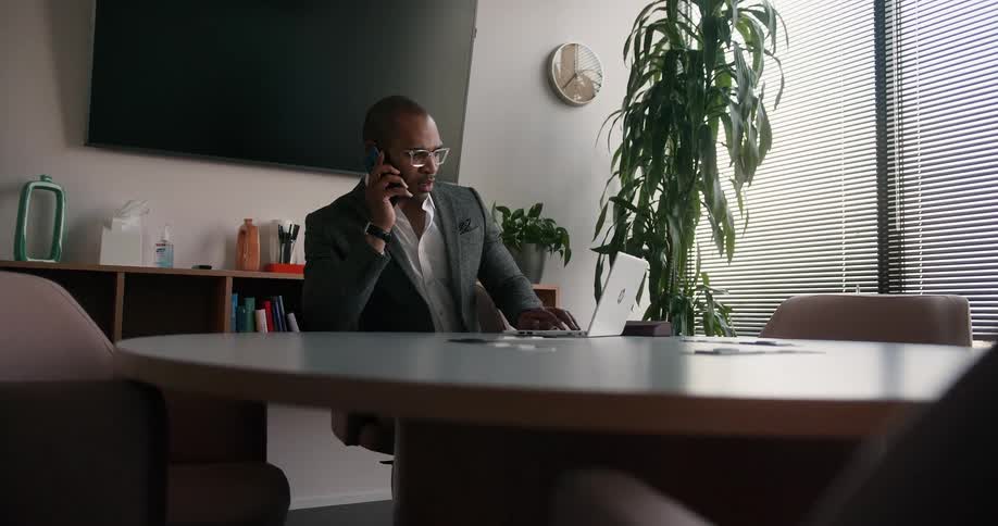 A man is sitting at a table with a laptop computer and he answers his phone