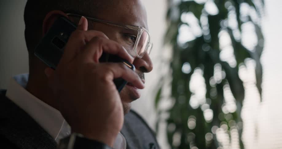 A man holding a smart phone up to his ear having a conversation near a window