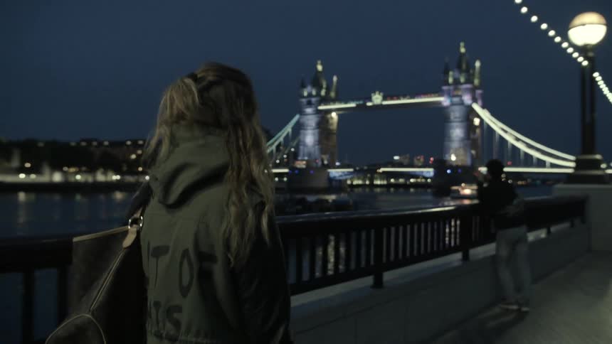 A woman walks along a river at night with buildings and a lit up bridge in the background