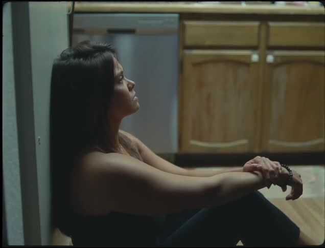 A teenage girl is leaning against a wall and she slides down to the floor where she sits and hits her head against the wall