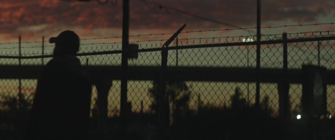 The silhouette of a man walking past a chain link fence with barbed wire on top