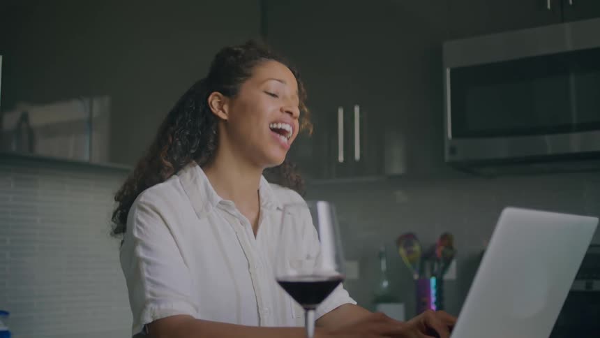 A woman is sitting down in front of a laptop computer laughing and talking to someone in a video chat