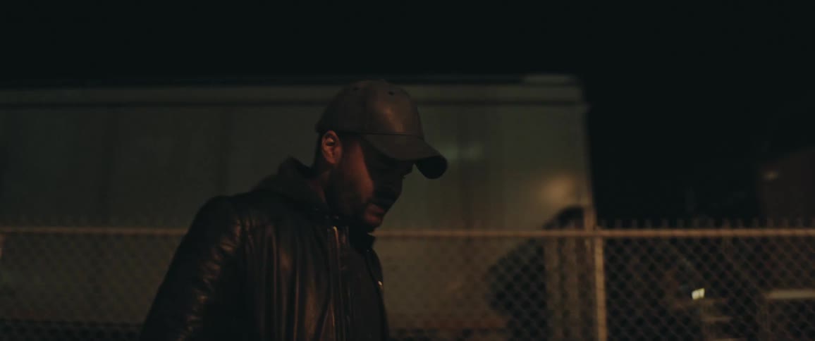 Man with cap beard and black leather jacket walks by fences and railway wagons at night
