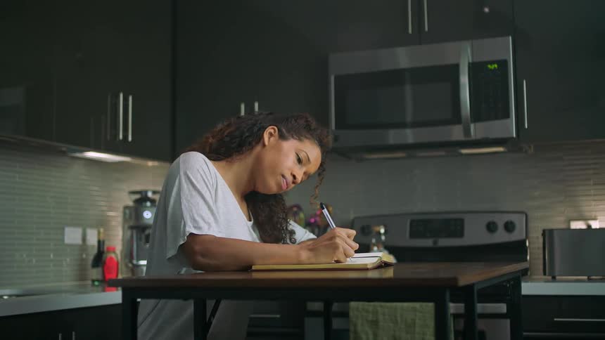 A woman writes in a journal while sitting at a small table in a kitchen