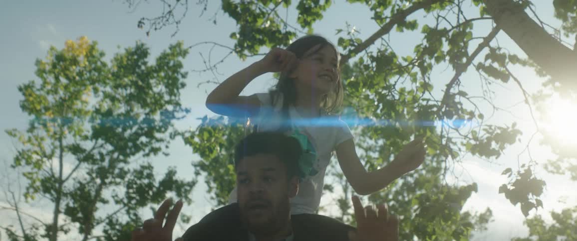 A man is outside dancing under a tree with his daughter on his shoulders as the sun shines brightly in the sky