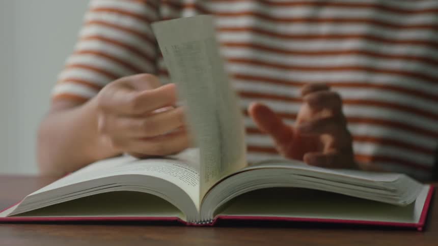 A girl wearing a striped shirt is sitting at a desk reading a book and turning the pages
