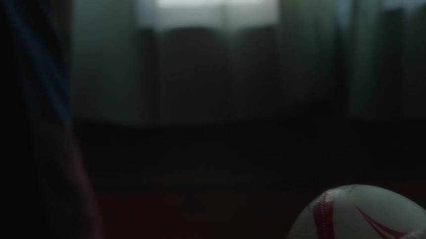 A boy sitting in his room holding a rugby ball turning it over in hands