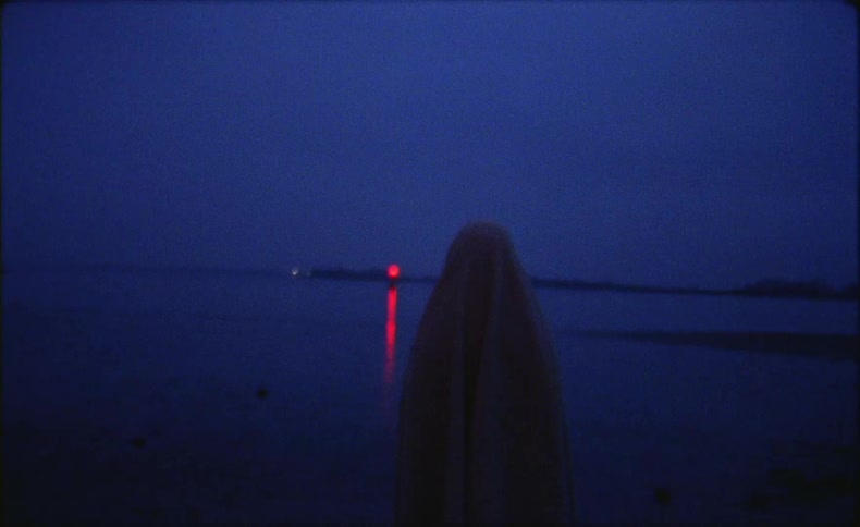 A figure draped in a sheet walking towards the water and a red flashing beacon