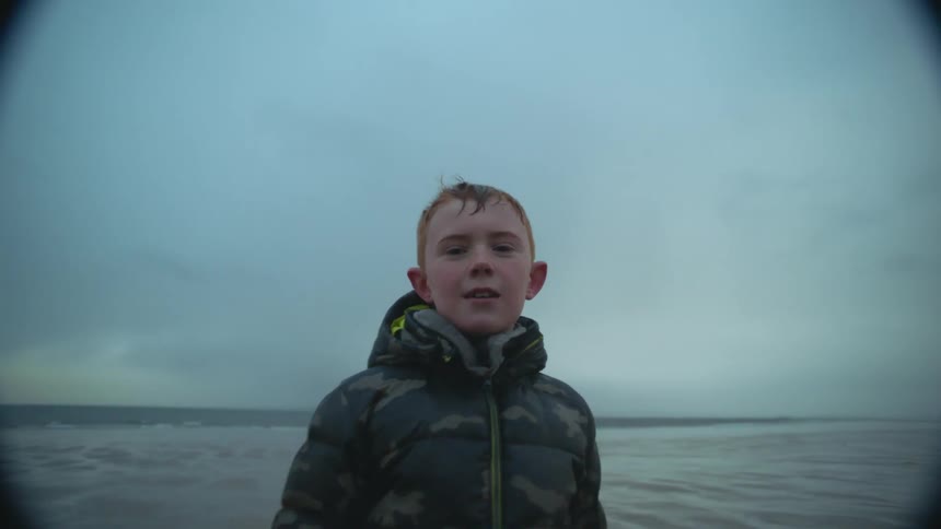 A boy walks along the coast in a puffer coat