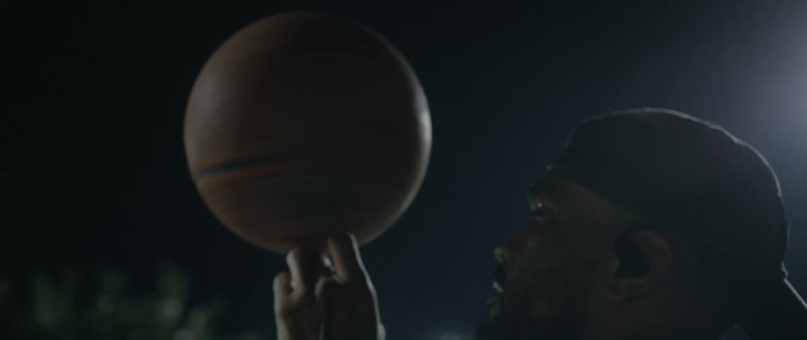 A man spins a basketball on his fingers while standing outside under the lights at night