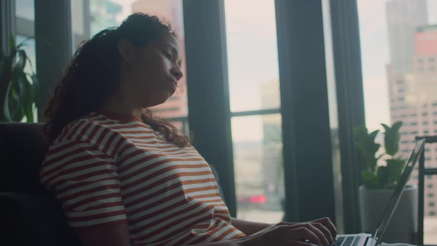 A woman sits in her home by a large window typing on a laptop