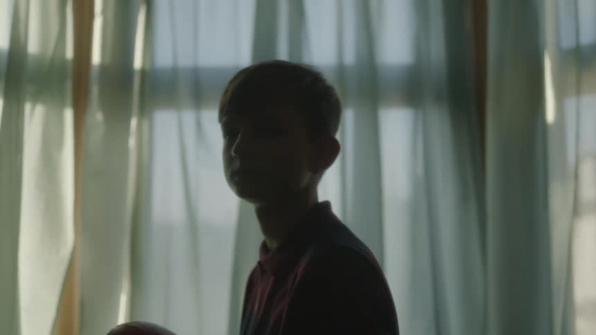 Boy holding football stands partly silhouetted near window in dark room