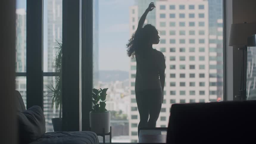 A woman is dancing in front of a window and there are high rise buildings outside
