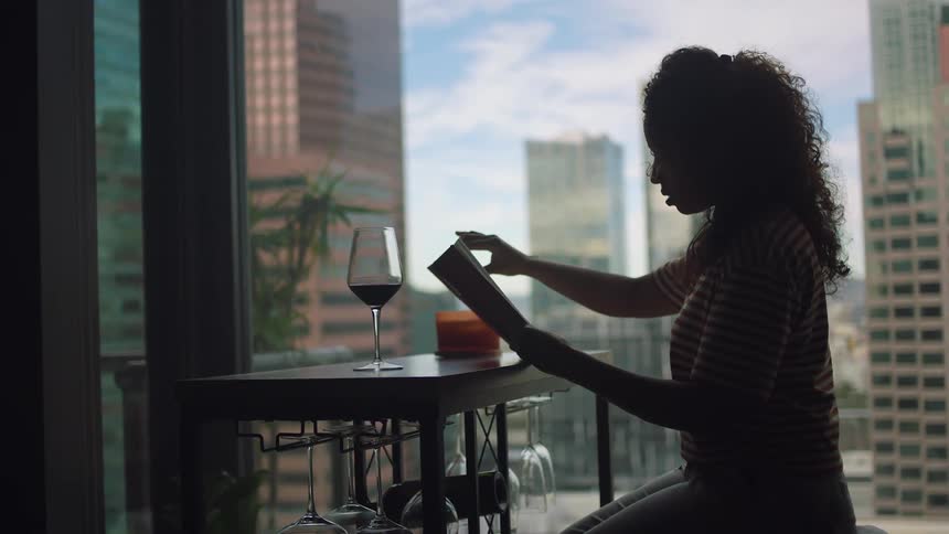 A woman sits at home and reads with a glass of wine