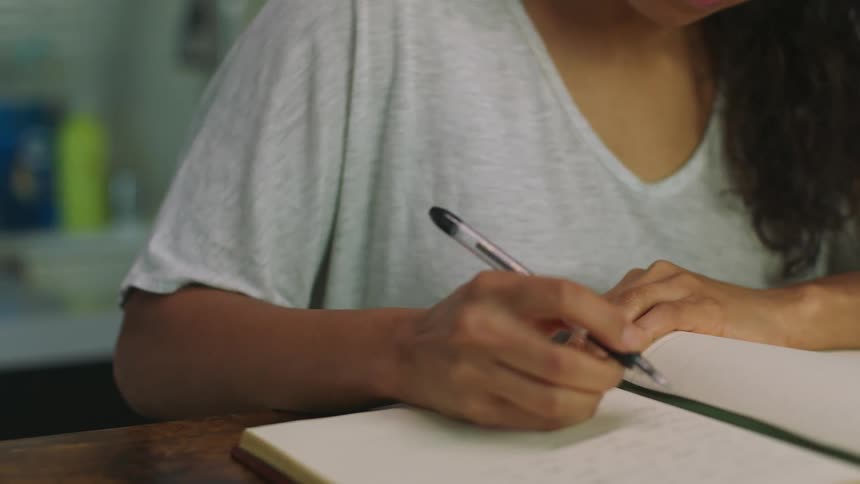 A woman sits at home and takes a drink of a hot beverage and then sets it down on a table and writes in a book