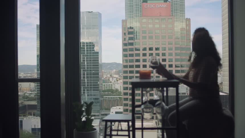 A woman is sitting at a table with a wine glass reading a book and skyscrapers can be seen through the window