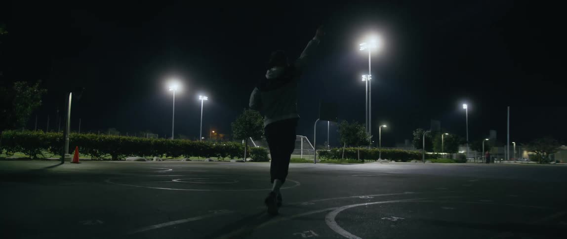A man carrying a basketball waves his hand at people playing on a court as he walks toward them